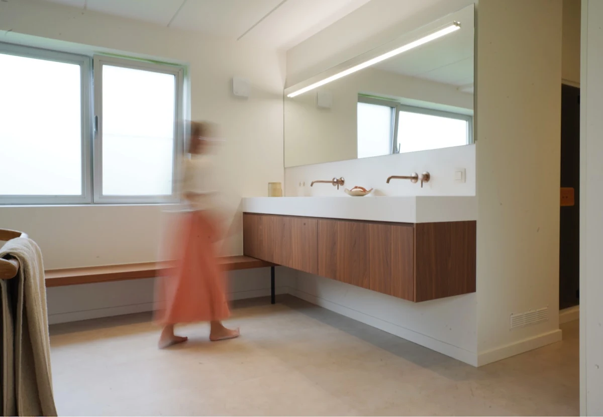 Lichte badkamer met houten badmeubel van muur tot muur, een vrouw wandelt voorbij